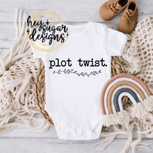 May include: White baby onesie with the text "plot twist." printed in black. The onesie is decorated with a small black floral design.