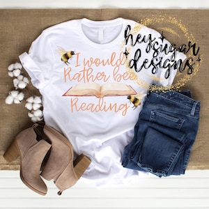 May include: White t-shirt with the text "I would rather be reading" and a book with bees on it. The shirt is on a brown surface with a pair of brown boots and blue jeans.