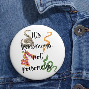 May include: White button with a black and white illustration of four snakes and the text "It's venomous, not poisonous".