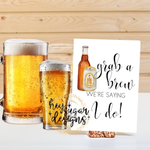 May include: White card with black text that reads "Grab a Brew We're Saying I Do!" with a watercolor illustration of a beer bottle and can. The card is propped up on a cork.