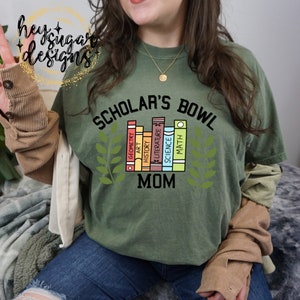 Può includere: Una maglietta verde con il testo "Scholar's Bowl Mom" e una grafica di una pila di libri con materie come geometria, arte, storia, letteratura, scienze e matematica.