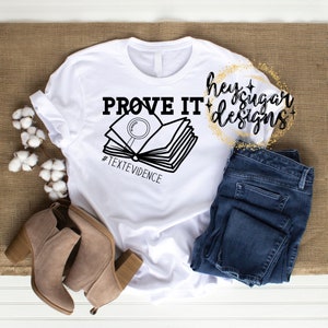 May include: White t-shirt with a black graphic of an open book with a magnifying glass on top. The text "PROVE IT" is above the book and "#TEXTEVIDENCE" is below the book. The text "hey sugar designs" is in a circle with gold glitter around it.