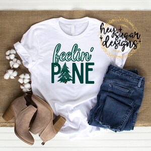 May include: White t-shirt with green text that reads "feelin' pine" and a green pine tree graphic.