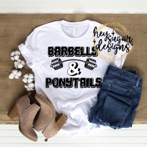 Op de afbeelding: Wit T-shirt met zwarte tekst die "Barbells & Ponytails" luidt met een halterstang afbeelding boven de tekst.