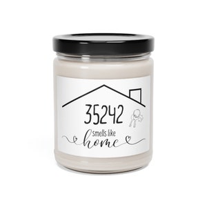 Puede incluir: Una vela perfumada en un frasco de vidrio transparente con tapa negra. La etiqueta presenta un contorno de casa, el número "35242" y la frase "smells like home" en escritura negra. También se incluye un gráfico de llave.
