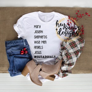 Puede incluir: Camiseta blanca con texto negro que dice "Mary, Joseph, Shepherds, Wise Men, Angels, Jesus, #SquadGoals". La camiseta está colocada sobre una superficie de arpillera marrón con un par de botines marrones de tobillo, pantalones vaqueros azules y una camisa a cuadros.