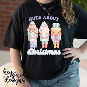May include: Black t-shirt with a graphic of three nutcracker figures in pastel colors. The text "Nuts About Christmas" is printed in purple and white.