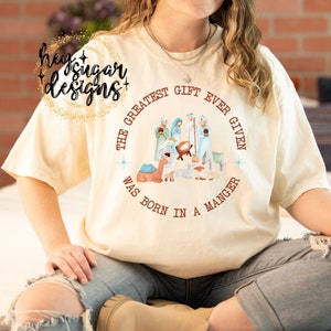 Puede incluir: Una camiseta beige con un diseño circular marrón que presenta una escena de la natividad y el texto "The Greatest Gift Ever Given Was Born In A Manger".