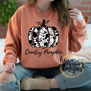 May include: A peach-colored sweatshirt with a black and white cow print pumpkin design and the text "Country Pumpkin".