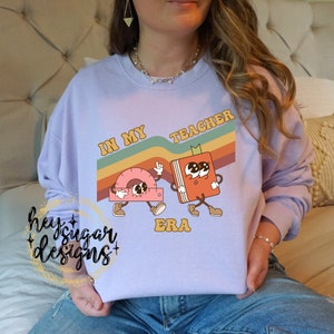 Puede incluir: Una sudadera de color lavanda con un diseño gráfico retro que presenta un libro y un transportador con el texto "In My Teacher Era".