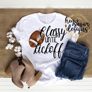 May include: White t-shirt with the text "Classy until Kickoff" and a watercolor football graphic. The shirt is paired with blue jeans and tan ankle boots. The image is on a neutral background.