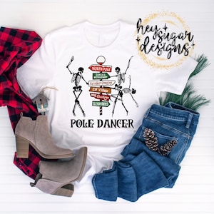 May include: White t-shirt featuring a graphic of a skeleton pole dancing with a sign that reads "North Pole, Santa's Workshop, Reindeer Crossing, Elf Village, Mrs. Claus Kitchen, Sleigh Rides". The text below the graphic reads "Pole Dancer".
