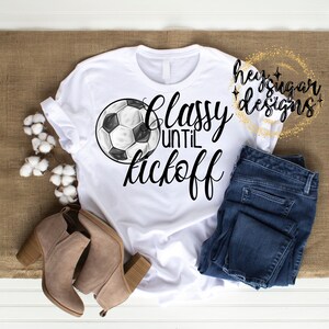 May include: White t-shirt with the text "Classy until kickoff" and a soccer ball graphic. The shirt is paired with blue jeans and tan ankle boots. The image also includes cotton stems.