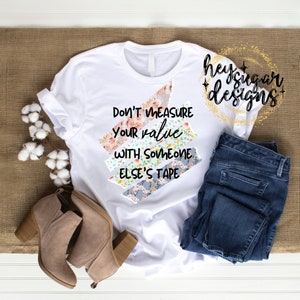 May include: White t-shirt with the slogan "Don't measure your value with someone else's tape." The text is layered over floral patterned tape strips. The shirt is paired with blue jeans and tan ankle boots. Cotton stems are on the left.