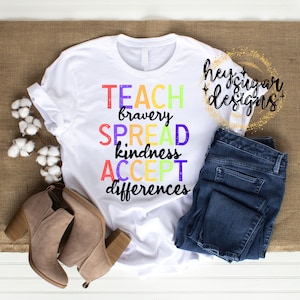 May include: White t-shirt with the text "TEACH bravery SPREAD kindness ACCEPT differences" in rainbow colors. The shirt is on a wooden surface with a pair of brown boots and blue jeans.