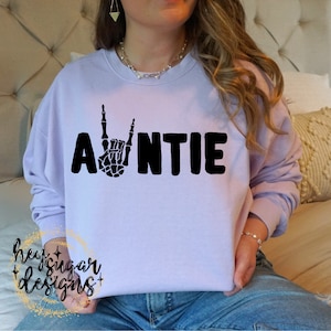 Puede incluir: Una sudadera de color lavanda con la palabra "AUNTIE" impresa en negro. La "A" está reemplazada por una mano de esqueleto haciendo el gesto de rock and roll.