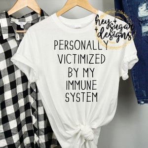 Puede incluir: Camiseta blanca con el texto "PERSONALLY VICTIMIZED BY MY IMMUNE SYSTEM" en negro. La camiseta está en una percha de madera. A la izquierda se ve una camisa a cuadros en blanco y negro.