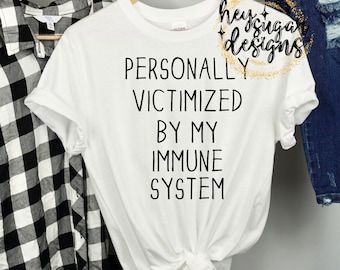 Peut inclure: T-shirt blanc avec le texte "PERSONALLY VICTIMIZED BY MY IMMUNE SYSTEM" en noir. Le t-shirt est sur un cintre en bois. Une chemise à carreaux noir et blanc est visible à gauche.