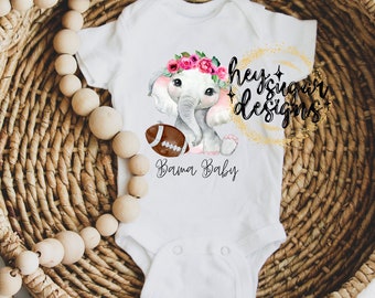 May include: White baby onesie with a watercolor illustration of a gray elephant wearing a pink flower crown and holding a brown football. The text "Hey Sugar Designs" and "Bama Baby" are printed on the onesie.