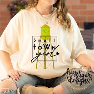 May include: A white t-shirt with a graphic of a water tower with the text "Billy Bob Loves Charlene" on it. The graphic is surrounded by a black square with the text "Small Town Girl" in black lettering. The text "hey sugar designs" is visible in the bottom right corner of the image.