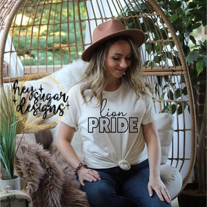 Puede incluir: Una camiseta blanca con el texto "lion PRIDE" en letras negras. La camiseta la lleva puesta una persona sentada en una silla de mimbre. El texto "hey sugar designs" es visible en el fondo.