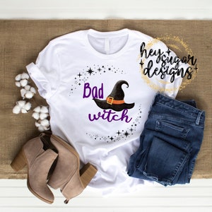 May include: White t-shirt featuring a purple and black witch hat graphic and the text "Bad Witch" surrounded by black stars. The shirt is on a brown surface with a pair of brown ankle boots and blue jeans.