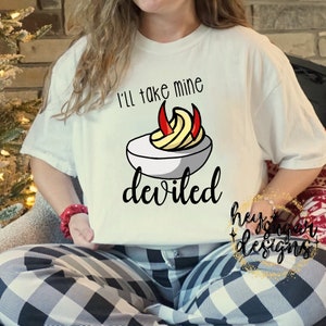 Könnte beinhalten: Weißes T-Shirt mit einem Grafikdruck eines teuflischen Eies mit roten Teufelshörnern und dem Text "Ich nehme meins teuflisch".