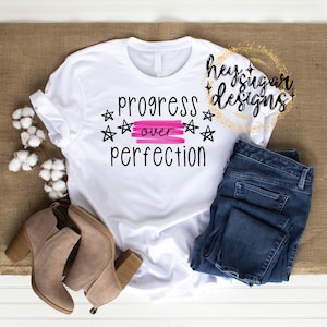 May include: White t-shirt with the text "progress over perfection" in black lettering with pink stars and a pink stripe. The shirt is laying on a brown surface with a pair of brown boots and blue jeans.