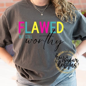 Op de afbeelding: Een grijs T-shirt met de tekst "FLAWED BUT STILL worthy" in kleurrijke letters. De tekst is in een vet, bloklettertype. Het shirt wordt door een persoon gedragen.