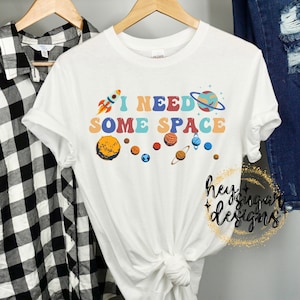 May include: White t-shirt with a graphic of a rocket ship, planets, and the text "I NEED SOME SPACE" in colorful retro lettering.