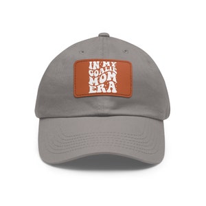 May include: A gray baseball cap with a brown patch that says "In My Goalie Mom Era" in white retro lettering.