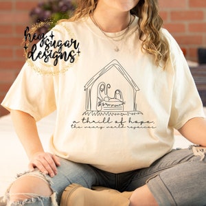 Puede incluir: Camiseta color crema con un dibujo lineal negro de un pesebre. El texto "a thrill of hope, the weary world rejoices" está debajo del dibujo. La camiseta tiene mangas cortas y un corte holgado. La camiseta se está usando.