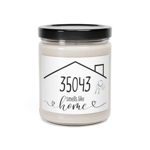Puede incluir: Una vela blanca en un tarro de vidrio con tapa negra. La etiqueta del tarro tiene una casa con el número 35043 y el texto "huele a hogar".