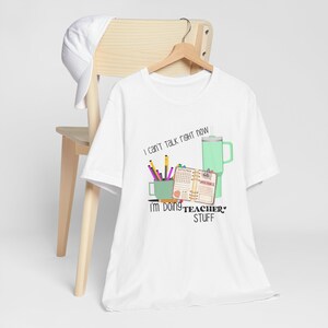 May include: White t-shirt featuring a graphic design of a coffee mug, pencils, a planner, and a tumbler. The text on the shirt reads "I can't talk right now I'm doing TEACHER STUFF".