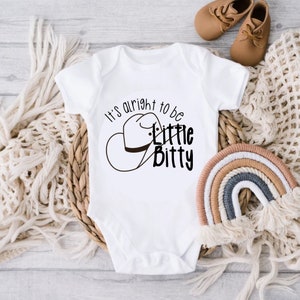 Peut inclure: Combinaison blanche pour bébé avec un graphique noir d'un chapeau de cowboy et le texte "It's alright to be little bitty".