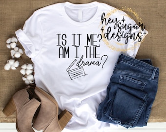 Könnte beinhalten: Weißes T-Shirt mit dem Text "Is it me? Am I the drama?" und einer Schwarzweißillustration einer Filmklappe. Das Shirt liegt auf einer braunen Jutestoffoberfläche mit einem Paar braunen Wildlederstiefeln und einer blauen Jeans.
