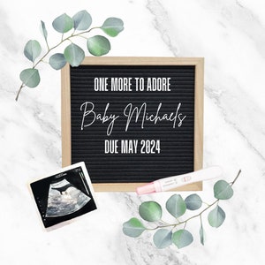 以下が含まれることがあります： 「One More to Adore Baby Michaels Due May 2024」と白文字で書かれた黒いレターボード。白黒の超音波画像とピンクの妊娠検査薬が、緑色のユーカリの葉が置かれた大理石の背景の上に置かれています。