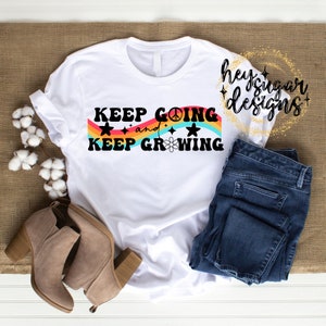 Puede incluir: Camiseta blanca con un gráfico de arcoíris y estrellas. El texto de la camiseta dice "Keep Going and Keep Growing".
