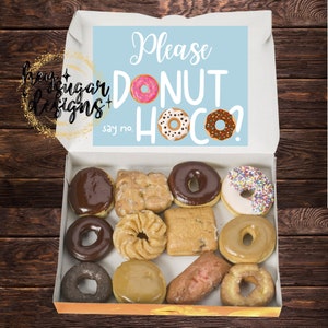 Puede incluir: Una caja blanca con una tapa azul que dice "Please donut hoco? say no." La caja está abierta y llena de una variedad de donuts, incluyendo donuts glaseados, de chocolate y con chispas.
