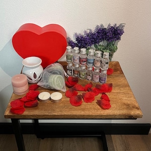 Op de afbeelding: Een houten tafel met een rode hartvormige doos, een witte keramische olieverdamper, een roze kaars, twee witte theelichtjes, een zakje groene wax melts, rode rozenblaadjes en een boeket lavendelbloemen met 15 kleine glazen flesjes met etherische oliën.