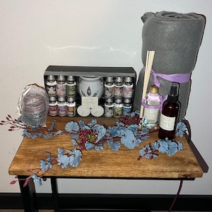 Op de afbeelding: Een houten tafel met een grijs deken, een set essentiële olieflessen, een diffuser, een rietdiffuser en een flesje kamerspray. De tafel is versierd met een blauwe en rode krans.