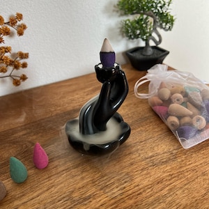Op de afbeelding: Zwarte keramische wierookhouder in de vorm van een hand die een paarse wierookkegel vasthoudt. De houder staat op een houten oppervlak met een klein zakje wierookkegels en een kleine bonsai op de achtergrond.