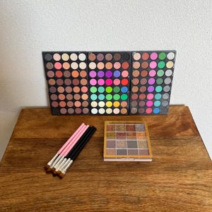 Op de afbeelding: Drie rechthoekige oogschaduwpaletten met een breed scala aan ronde pans in neutrale, levendige en glitterende tinten. Daarnaast liggen vier make-upkwasten met roze en zwarte handvatten en een vierkant palet met glitterpigmenten.