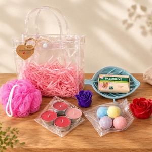 Op de afbeelding: Een doorzichtige plastic geschenktas gevuld met roze papiersnippers en een hartvormig label, een felroze badspons, vier rode theelichtjes, een blauw bladvormig zeepbakje met een Palmolive zeepblok en vier kleine bruisballen voor in bad.