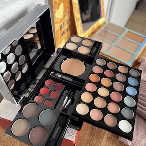 Op de afbeelding: Een make-up paletset met meerdere lagen, bestaande uit een grote zwarte opvouwbare cassette met talloze ronde oogschaduws in neutrale, metallic en pastelkleuren, rode lippenkleuren, een poeder, twee kleine kwastjes en een wafel-thema palet.