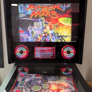 May include: A pinball machine with a red and black cabinet. The backglass features a colorful image of aliens and a flying saucer with the text "Attack from Mars". The game is lit up and ready to play.