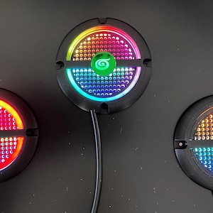 May include: Three black, circular LED lights with colorful, pixelated patterns. Each light has a central button and a wire. The lights display red, orange, yellow, green, blue, and purple hues.