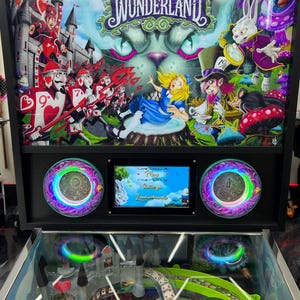 May include: A pinball machine with an "Alice Goes to Wonderland" theme. The artwork features Alice, the Cheshire Cat, the Mad Hatter, and the White Rabbit. The machine has a screen displaying game options and two circular light displays.
