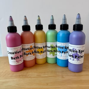 May include: Six squeeze bottles of Star Bright Body Care Creative Spark Bath Paint in pink, orange, yellow, green, blue, and purple. Each 4 oz bottle features a black cap, a white label with a paint splatter graphic, and scent text.