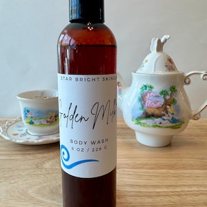 Puede incluir: Una botella de gel de ducha marrón con tapa negra y etiqueta blanca que dice "Golden Milk" y "Body Wash 8 OZ / 226 G". Una taza de té y una tetera con diseños florales están en el fondo.